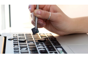 A woman's hand holding a key with tweezers above a laptop keyboard with numerous missing keys.